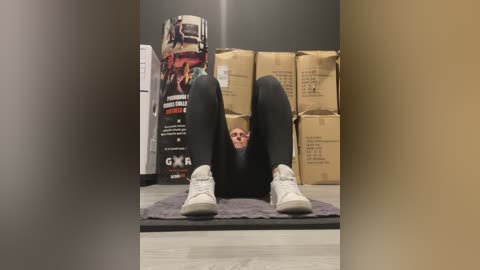 Media: Video of a person with short blonde hair, wearing black leggings and white sneakers, lying on a grey rug with legs up, in a storage room with brown cardboard boxes and a large poster.