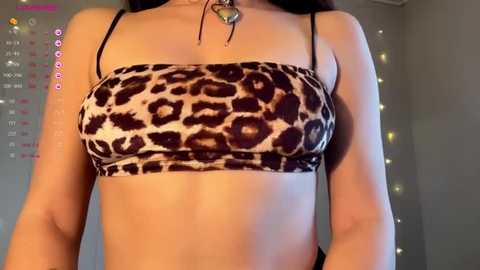 Video of a woman with fair skin wearing a leopard print bandeau top, black bra straps visible, with a heart necklace. Background features a calendar and string lights.