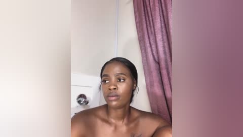 Media: Video of a bare-chested Black woman with dark skin, short hair, and a neutral expression, standing in a bathroom with beige walls, a white door, and maroon curtains.