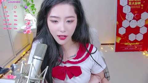 Media: Video of a young Asian woman with long black hair, wearing a red and white sailor uniform, singing into a microphone, surrounded by a colorful, cluttered desk and a red poster in the background.