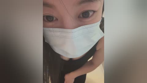 Media: Video of an Asian woman with a light complexion and long, straight dark hair, wearing a white surgical mask, black top, and pants, standing in a dimly lit room.