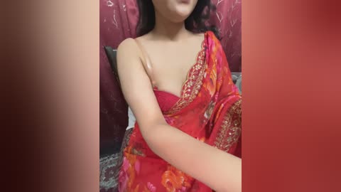 Media: Video of an Asian woman with light skin, wearing a vibrant red saree with gold embroidery, reclining against a patterned red background.