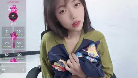 Media: A video of an East Asian woman with straight, shoulder-length black hair, wearing a green and navy blue hoodie with cartoon characters, sitting in a chair, with a virtual pregnancy app overlay showing 58 days.