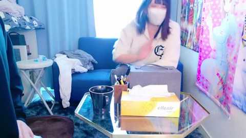 Media: Video of a woman in a white shirt and mask, sitting on a blue couch in a cluttered room with blue curtains, a colorful poster, and a glass table with tissues and a box.