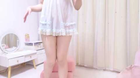 Media: A video of a fair-skinned woman in a sheer, white babydoll lingerie with light blue trim, standing in a pastel-colored bedroom with a vanity table, mirror, and pink ottoman.