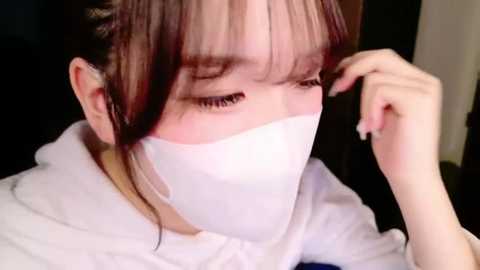 Media: Video of a young woman with fair skin and dark hair, wearing a white face mask, adjusting it with her right hand. She has bangs and is dressed in a white robe. The background is dark and indistinct.