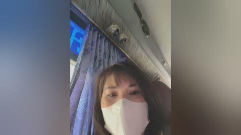 Media: Video of a young Asian woman with light skin, wearing a white surgical mask, brown hair, and a dark jacket. She is in a plane, with a blue curtain and overhead lights visible.