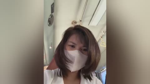 Media: Video of an East Asian woman with straight, shoulder-length brown hair, wearing a white surgical mask, standing in a bathroom with a mirror, green walls, and a towel rack.