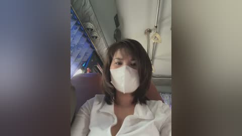 Media: Video of a woman with medium skin tone, shoulder-length dark hair, wearing a white face mask and white shirt, seated in an airplane with a grey wall and visible window.