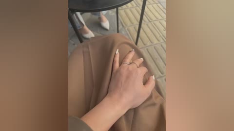 Media: A close-up video of a hand with manicured nails, wearing a brown ring, resting on a beige cloth over a person's lap. The background shows a tiled floor and a black chair.