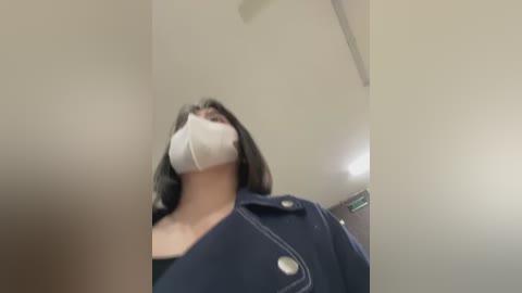 Media: Video of a woman with shoulder-length dark hair, wearing a white mask and navy jacket, taken from a low angle, with a blurred background.