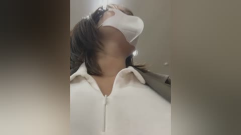 Media: Video of a person wearing a white surgical mask over their nose and mouth, with their head tilted back, revealing a white polo shirt with a zipper. The background is blurred and neutral-toned, creating a focused and clinical atmosphere.