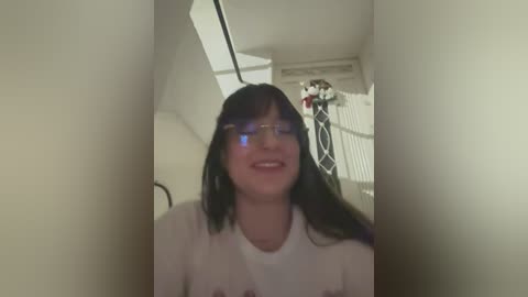 Video of a smiling, fair-skinned woman with long black hair and glasses, wearing a white t-shirt, standing in a dimly lit room with a window, blurred background, and a cat.