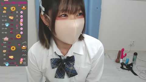 Media: Video of an Asian woman in a white shirt and blue plaid bow tie, wearing a surgical mask, with a white background and a blue curtain.