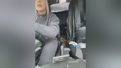 Media: Video of a blonde woman in a grey hoodie driving, with a messy car interior, including a leather jacket, snacks, and a water bottle.