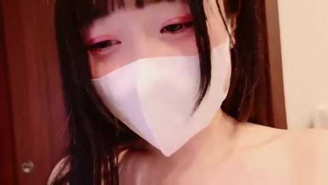 Media: Video of an Asian woman with long black hair and bangs, wearing a white face mask, with pink eyeshadow, in a dimly lit room with a wooden door.