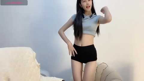 Media: Video of an East Asian woman with long black hair, fair skin, wearing a light gray crop top and black shorts, flexing her bicep, in a simple, light-colored room.