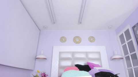 Media: Video of a pastel-colored bedroom with lavender walls, a white headboard, and a white window with grid pattern. Bed is cluttered with colorful pillows in pink, purple, and green. Decorative round wall sconces hang on either side of the headboard.