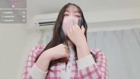 Media: Video of an Asian woman with long black hair, wearing a pink checkered sweater and a pink mask, standing indoors with an air conditioner and white curtains in the background.