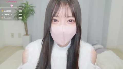 Media: Video of an East Asian woman with long, straight black hair and pale skin, wearing a white mask, in a minimalist, white room with a plant in the background.