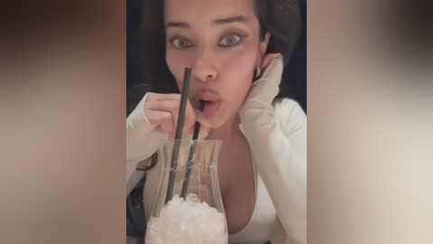 Video of a woman with long black hair, wearing a white top, drinking from a glass with a straw, looking surprised.