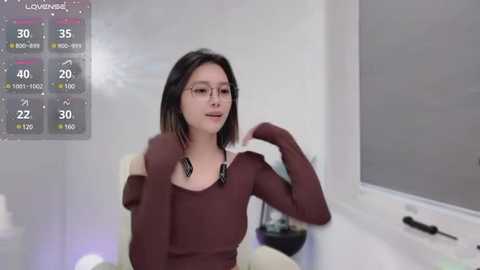 Media: Video of a woman with straight black hair, wearing a dark brown off-the-shoulder top, posing in a modern bathroom with a digital shower display and window blinds.