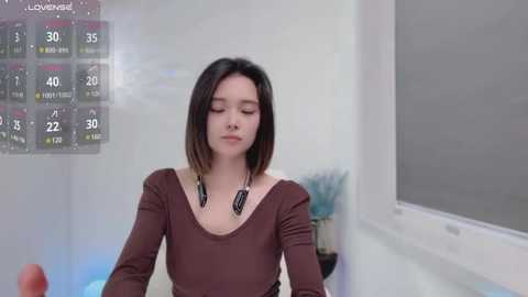 Media: Video of a young Asian woman with straight, shoulder-length dark hair, wearing a brown long-sleeve top, standing in a modern hallway with a digital display showing temperature and humidity levels.