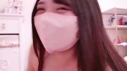 Media: Video of a young Asian woman with long black hair, wearing a white surgical mask, in a bright, cluttered room with white shelves and colorful toys.