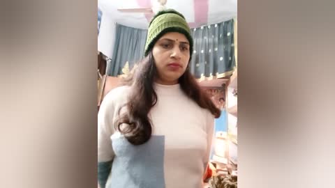 Media: Video of a South Asian woman in a beige sweater, green beanie, and light makeup, looking pensive in a cluttered, dimly lit room with pastel walls and a wooden dresser.