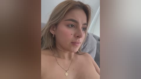 Media: Video of a young woman with light skin, shoulder-length blonde hair, wearing hoop earrings and a gold necklace, sitting topless on a gray couch, looking contemplative.