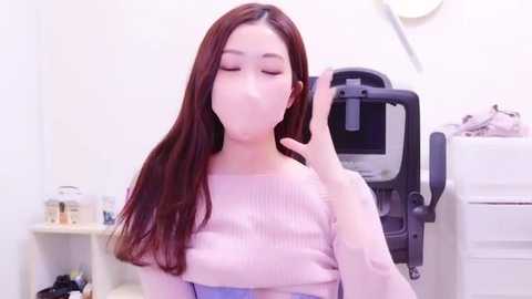 Media: Video of an Asian woman with long, straight brown hair, wearing a light pink ribbed sweater, sitting in a brightly lit room with white walls. She's blowing a bubble with a satisfied expression.