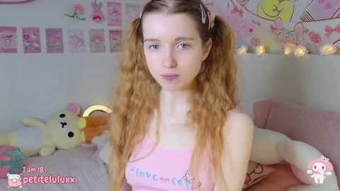 Media: A video of a young woman with long, curly blonde hair in pigtails, wearing a pink tank top with \"love\" written on it, sitting on a bed with stuffed animals and pink decorations in the background.