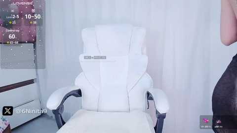 Media: Video of a white gaming chair in a minimalist room with a white curtain background. A person in a dark shirt is partially visible on the right.