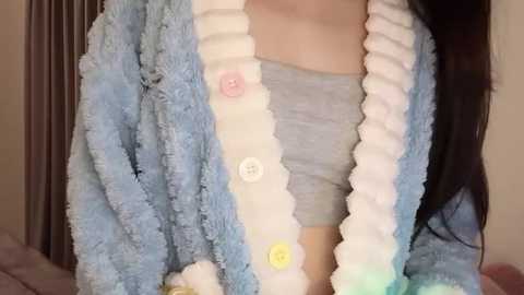 Media: Video of a person wearing a fluffy, light blue cardigan with white trim, featuring large buttons, over a light gray shirt. The background includes a dark curtain and a soft, beige wall.