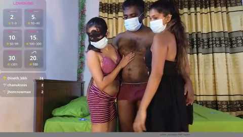 Media: A video captures a man in a hospital gown, wearing a face mask, standing between two women in lingerie, one in a red bra and skirt, the other in a black dress. The background features green curtains and a bed.