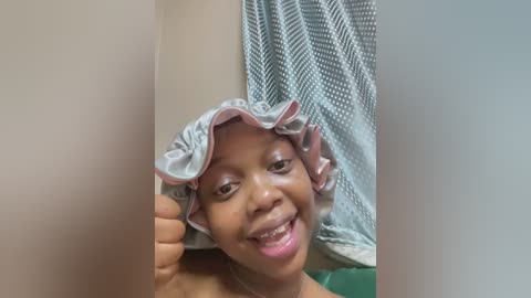 Media: A video of a young Black girl with a light brown complexion, smiling while holding a wet, folded towel on her head, in a bathroom with a blue shower curtain.