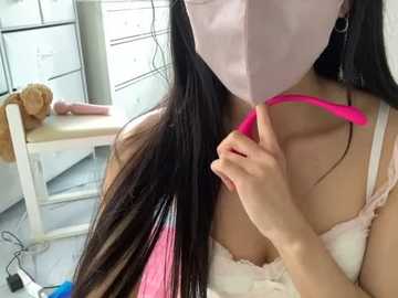 Media: Video of an East Asian woman with long, straight black hair, wearing a pink face mask and a white lace bra, holding a pink vibrator, seated in a white room with a wooden chair and a stuffed teddy bear.