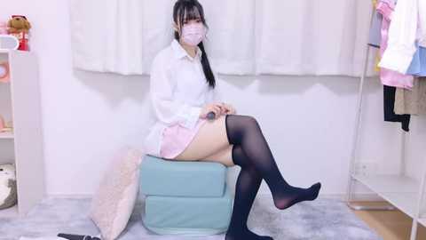 Media: A video of an Asian woman with long black hair, wearing a white mask, white blouse, pink skirt, and black thigh-high stockings, sitting on a blue cushion in a simple room with white walls and clothing racks.