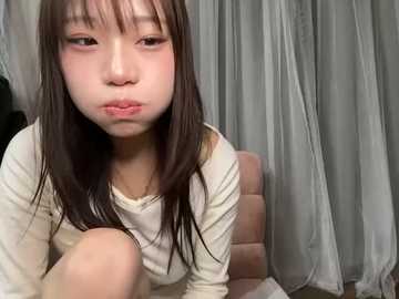 Media: Video of a young Asian woman with straight, dark brown hair, wearing a white long-sleeved top, sitting on a chair. She has a pensive expression. Background features a curtain and a dark object on the left.