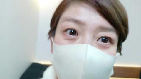 Media: Video of a young Asian woman with light skin and brown hair, wearing a white surgical mask that covers her mouth and nose, looking up with wide, expressive eyes.