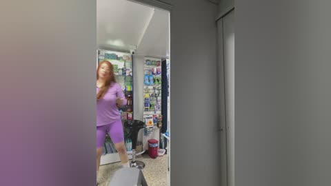 A video showing a young woman with long red hair in a purple nurse outfit, standing in a doorway. Behind her, a cluttered room with a fridge, shelves, and a red chair.