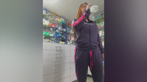 Video of a woman in a purple tracksuit, drinking water in a pharmacy with shelves full of colorful medicines in the background.