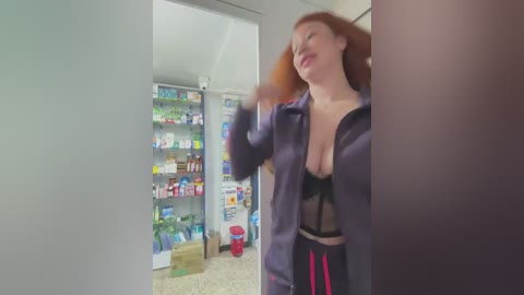 Media: Video of a red-haired woman in a purple jacket, exposing a black lace bra, dancing in a brightly lit pharmacy.