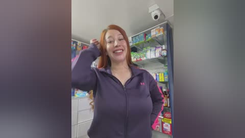 Media: Video of a smiling, fair-skinned woman with long red hair, wearing a purple zip-up jacket with pink accents, standing in a brightly lit pharmacy. Shelves behind her are stocked with various products.