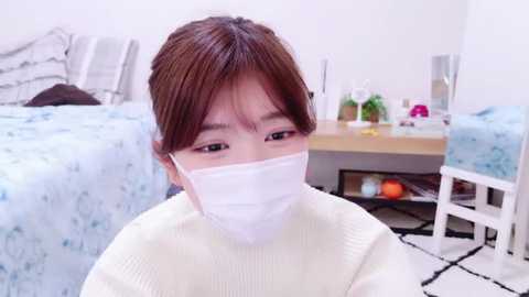 Media: Video of a young Asian woman with shoulder-length brown hair, wearing a white face mask, white sweater, and white earphones. She sits on a black-and-white patterned rug in a light-colored bedroom with a bed and white furniture.