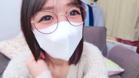 Media: Video of an Asian woman with straight, shoulder-length black hair, wearing round glasses, a white face mask, and a cream-colored knitted sweater. She sits on a gray couch with colorful pillows, in a room with a yellow curtain.