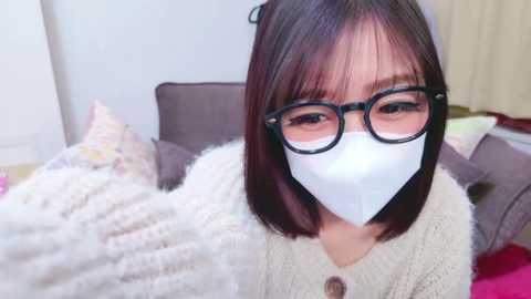 Media: Video of an East Asian woman with straight, shoulder-length dark hair, wearing black-framed glasses and a white face mask, sitting on a bed with a beige blanket and pillows.