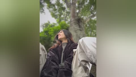 Media: A video of a young woman with long dark hair, wearing a brown coat, sitting on a bed in a forest, looking upwards, surrounded by green foliage.