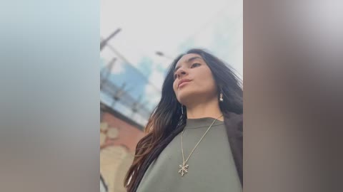 Media: A video of a young woman with long, dark hair, wearing a green top and a silver cross necklace, standing outdoors against a blurry urban background.