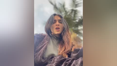 Media: Video of a young woman with long brown hair, wearing a light green sweater and a dark jacket, standing outdoors against a blurred background of greenery. She appears emotional, mouth slightly open.